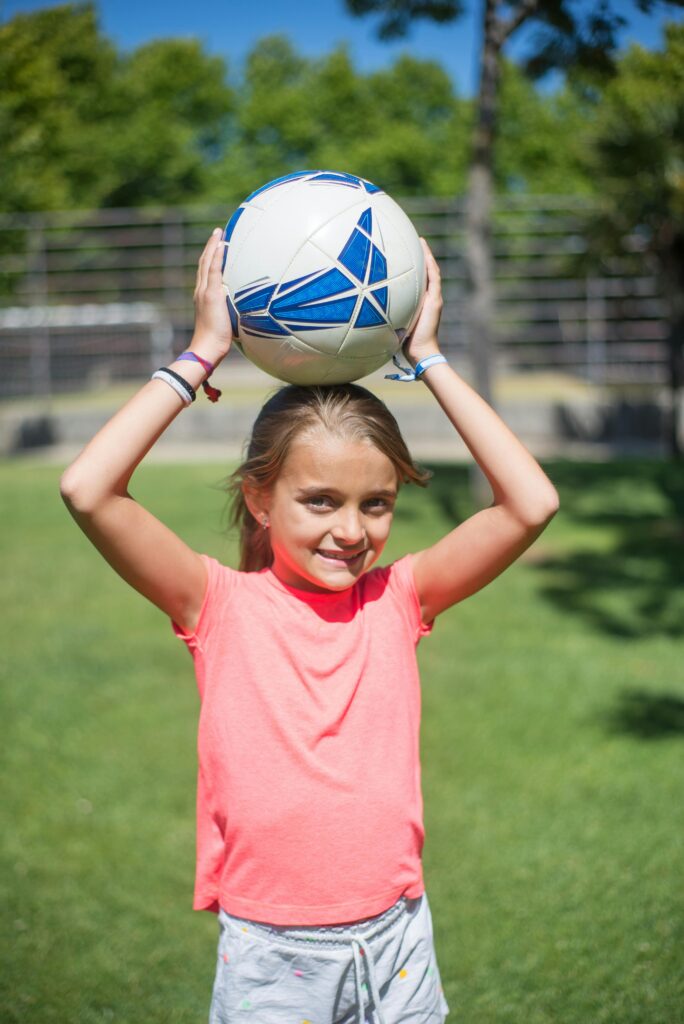 Smiling girl holding a soccer ball above her head on a sunny day outdoors.